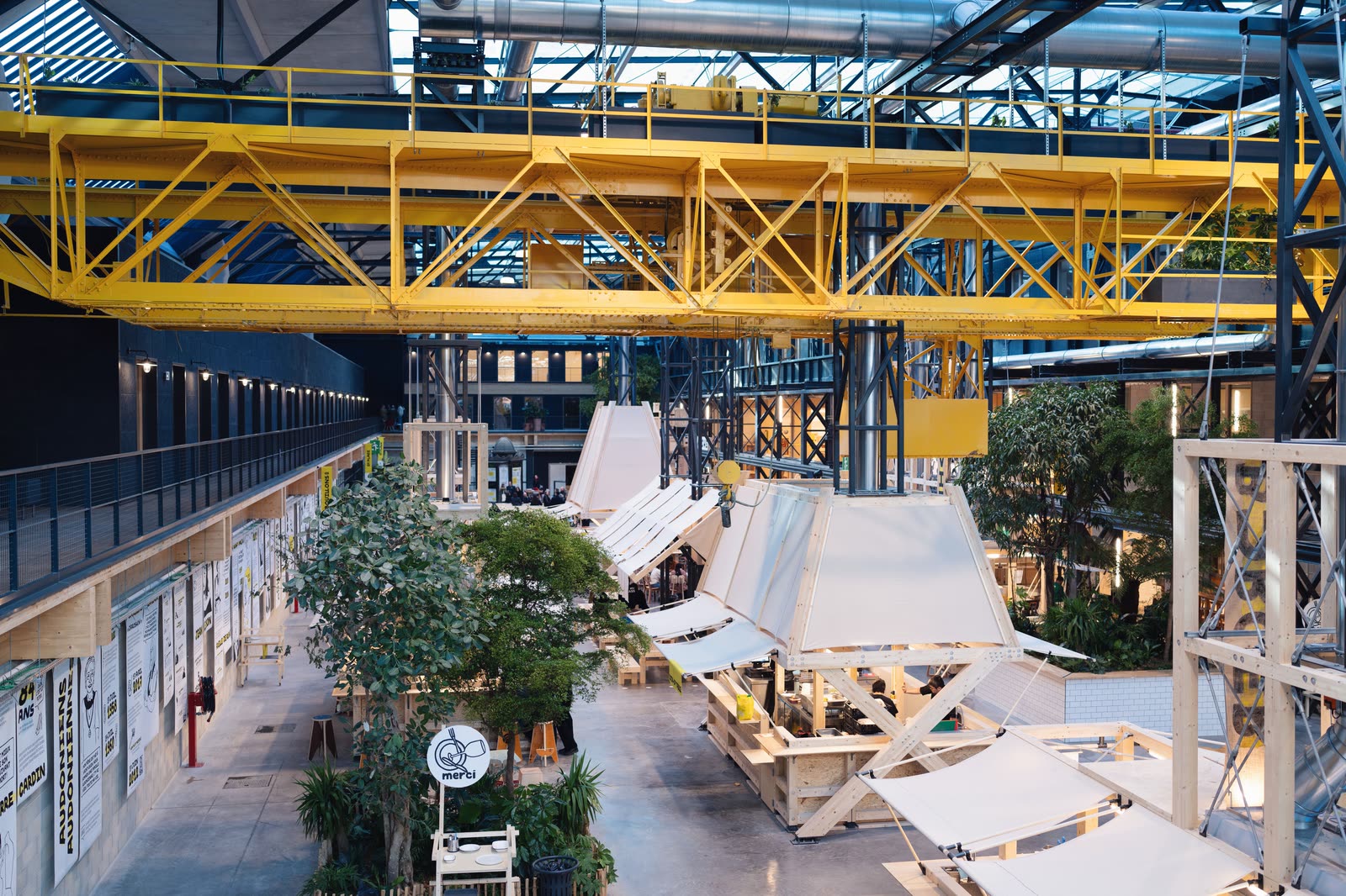 Inside the Communale, the food hall in St Ouen where KALIMERA has its restaurant: a yellow steel gantry spans the hall above white-roofed counters and trees