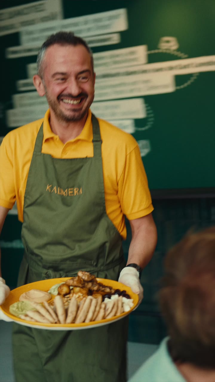 A smiling member of the KALIMERA team, in a yellow polo and olive apron, carrying a plate of Greek food to the table