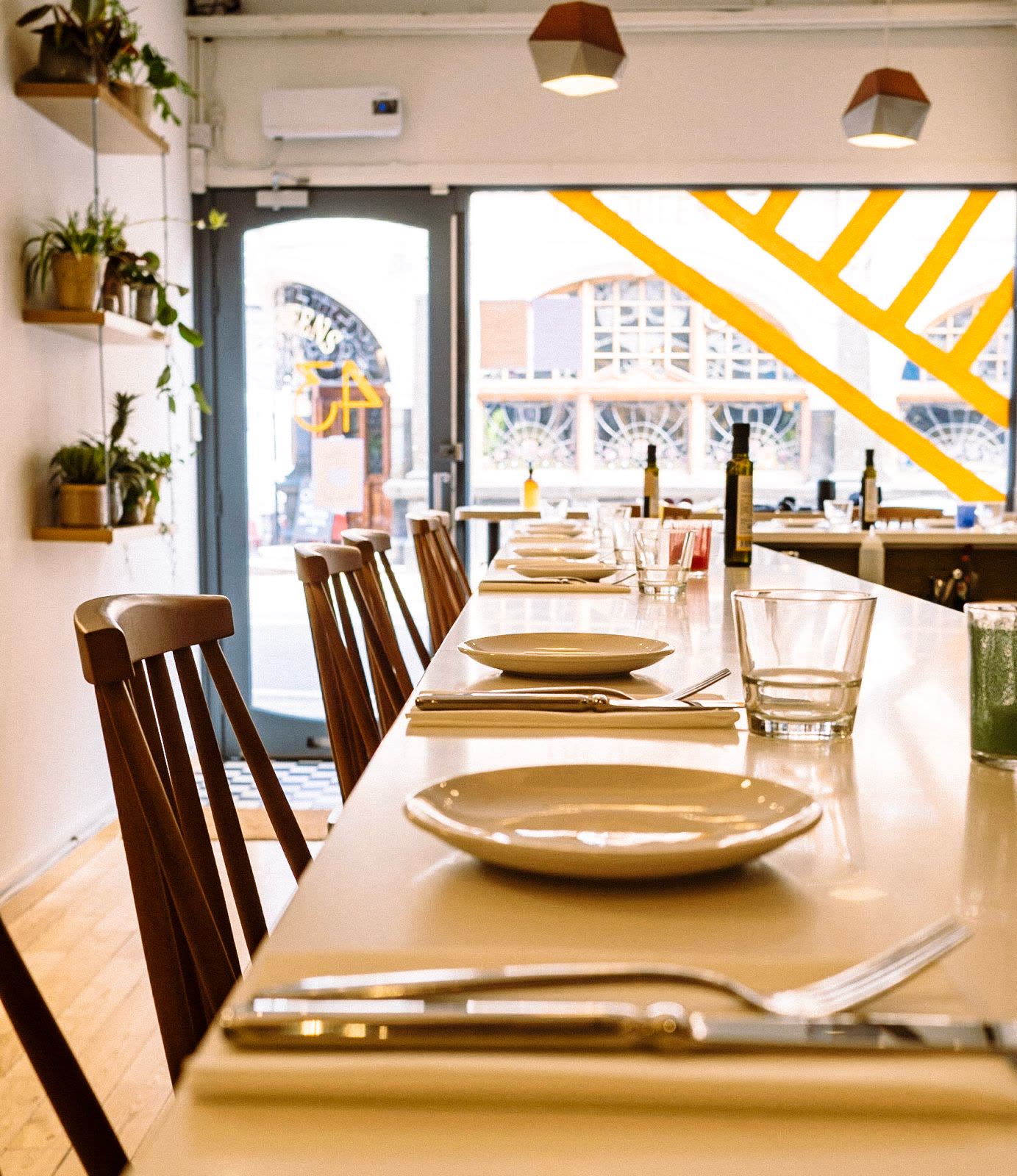 Inside KALIMERA in Crouch End, London: a long white counter laid with plates, cutlery and glasses, wooden chairs along it, plants on the wall and the yellow stripes of the shopfront across the window