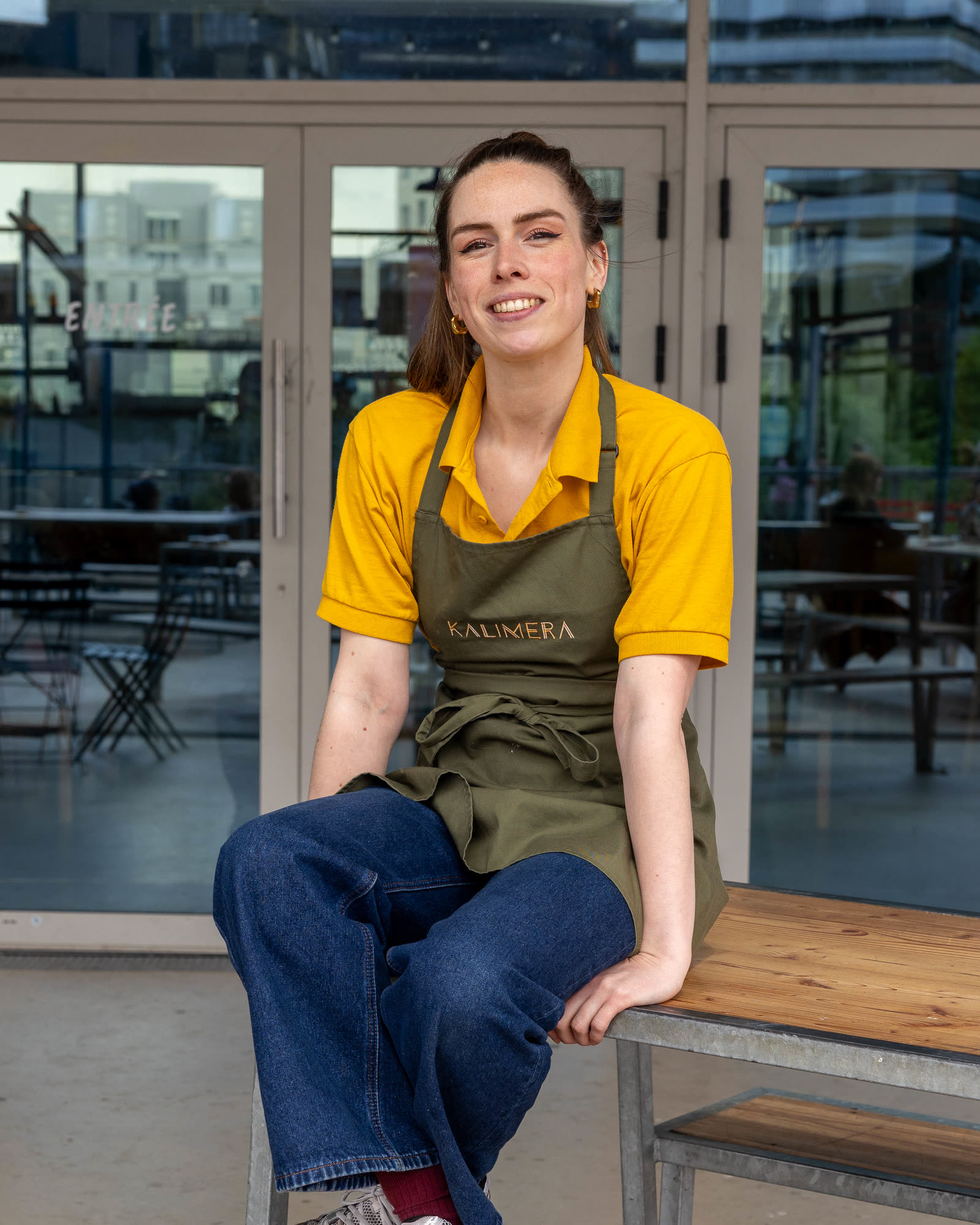 Une membre de l’équipe KALIMERA, polo jaune et tablier olive, tout sourire, assise sur un banc en bois