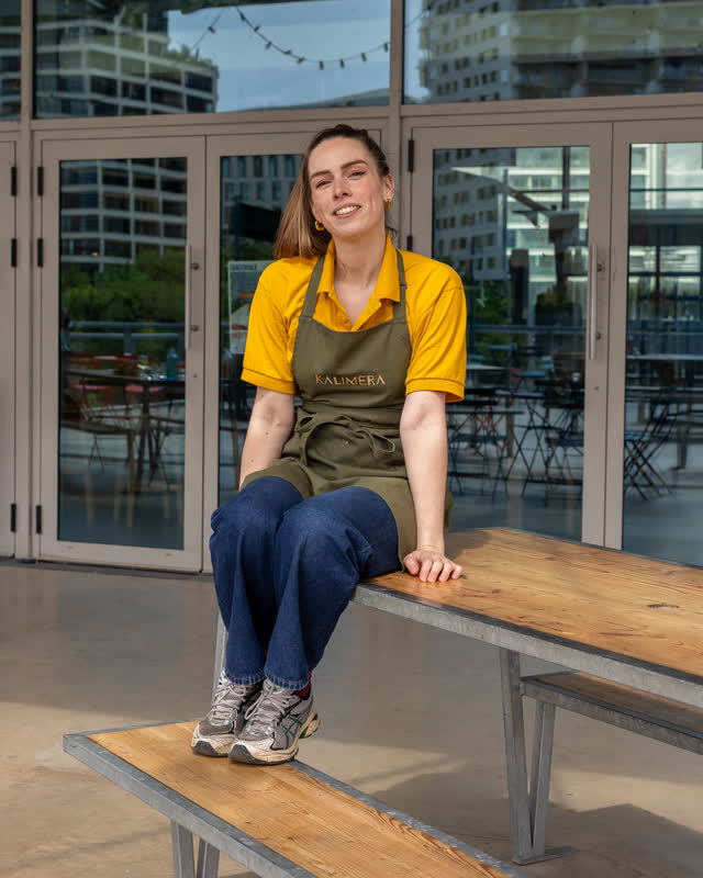 A KALIMERA team member in a yellow polo and olive apron sitting on a wooden bench, laughing