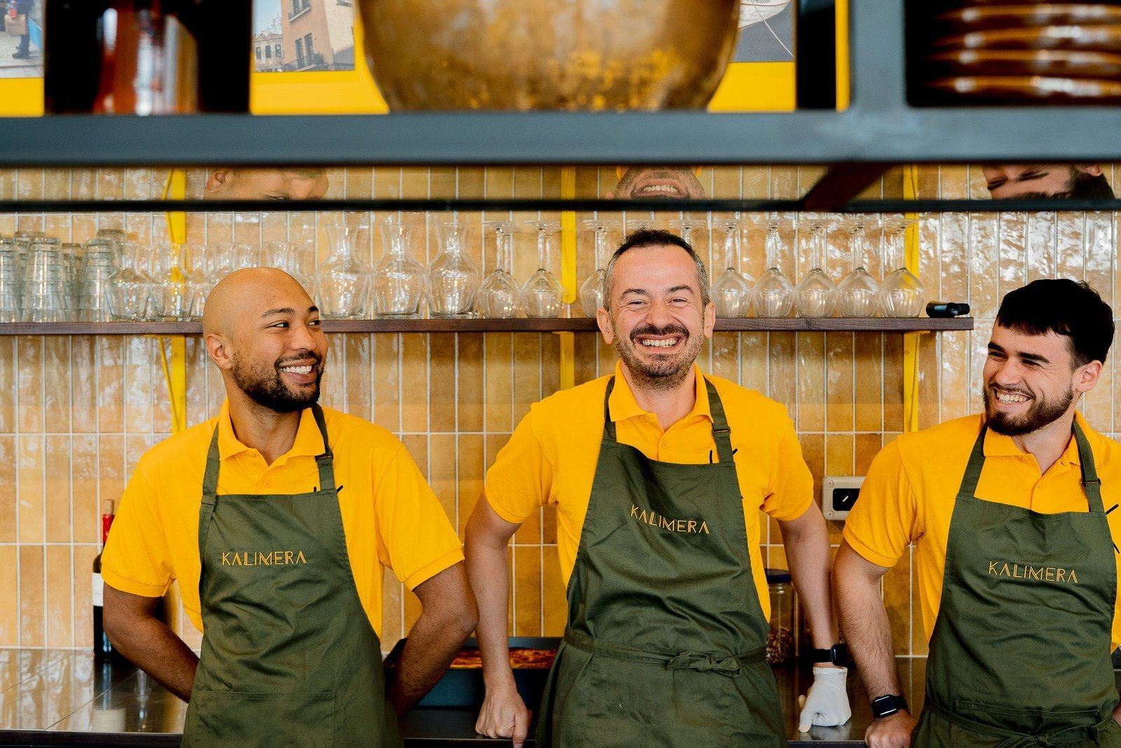 Trois membres de l’équipe KALIMERA rient derrière le comptoir, en polo jaune et tablier olive