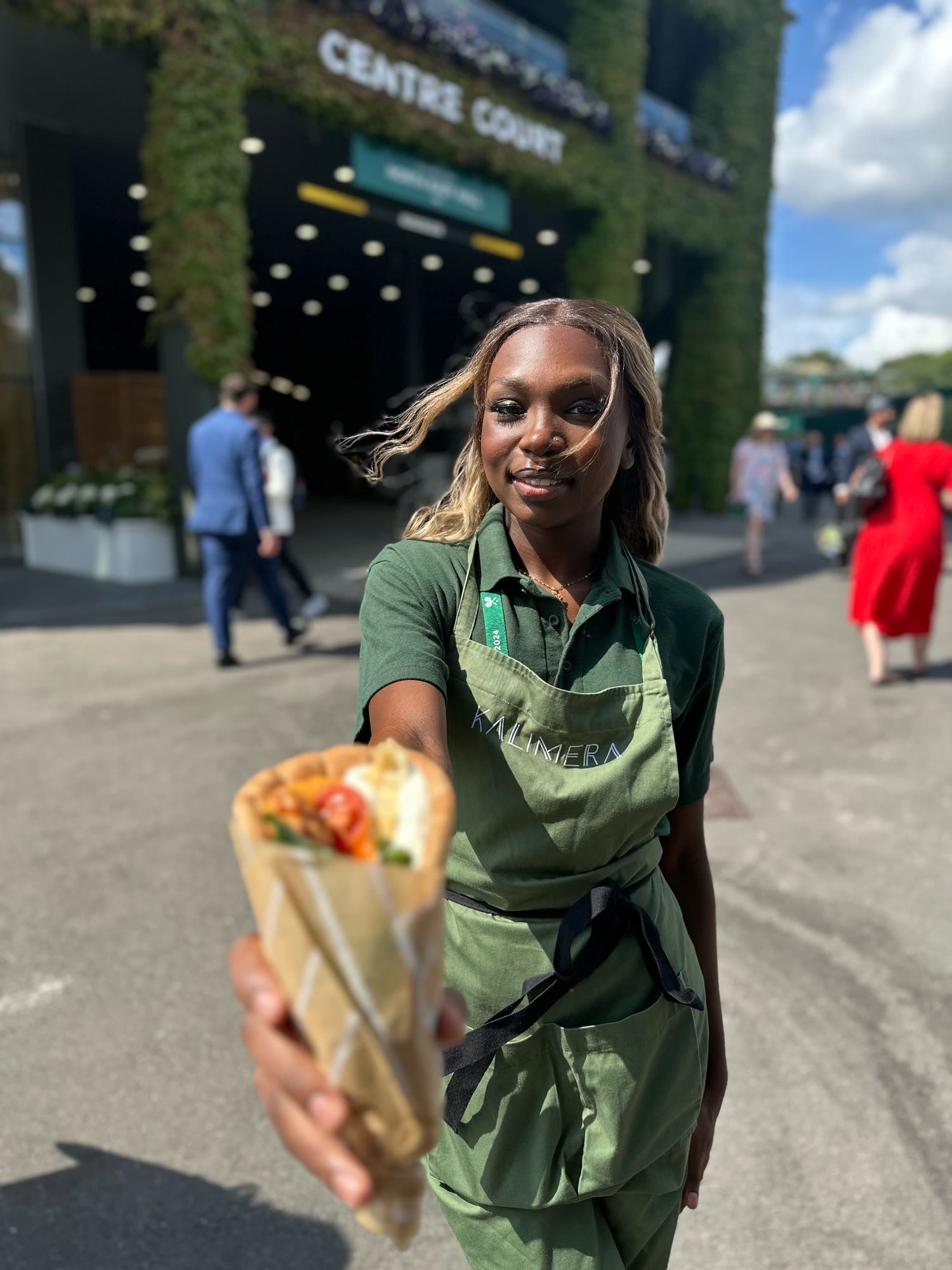 Une membre de l’équipe KALIMERA, tablier olive, tend un wrap devant le Centre Court de Wimbledon