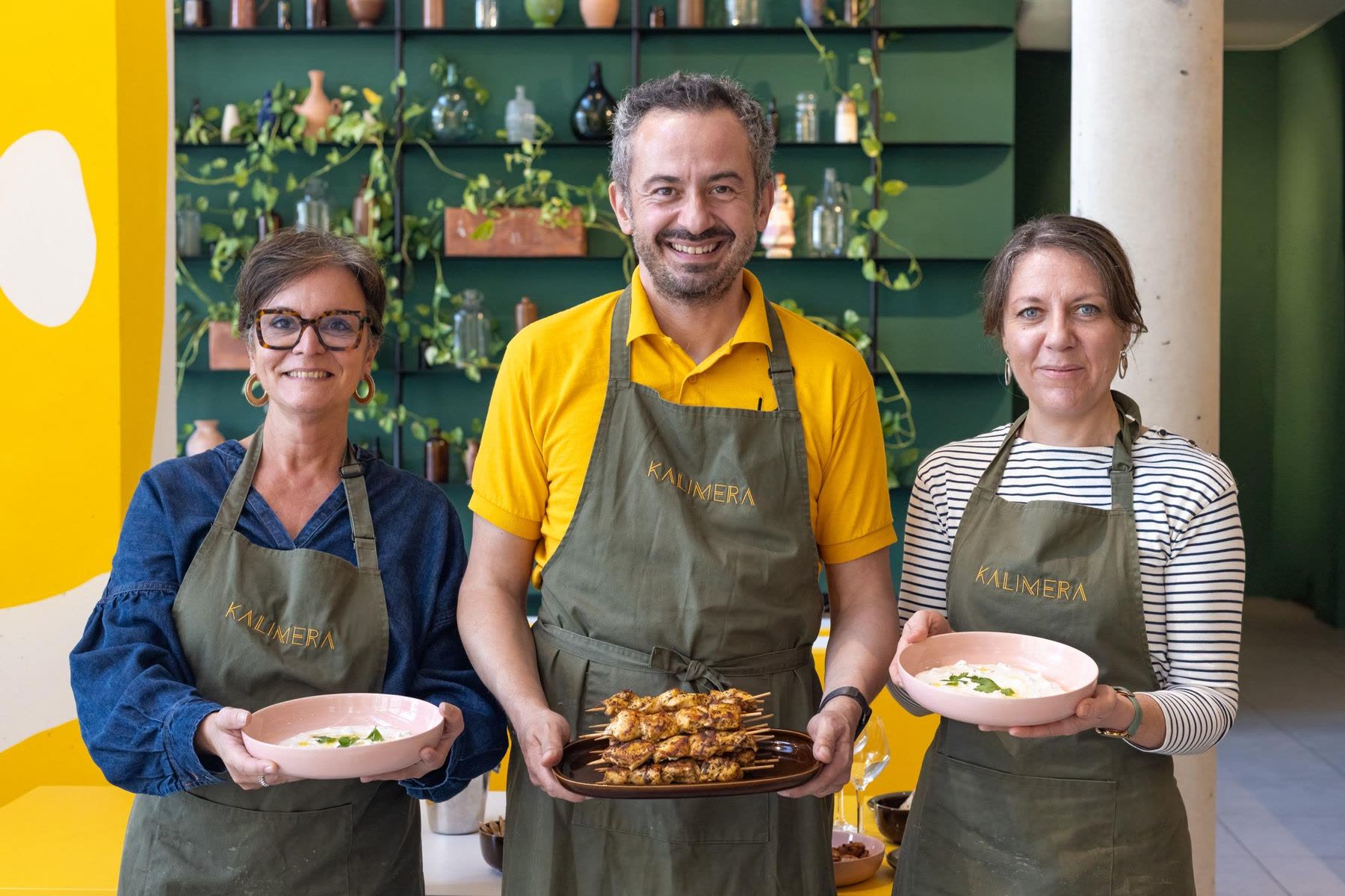 After a KALIMERA cooking workshop in Bordeaux: two guests and their host, in olive aprons, hold up the tzatziki and the chicken skewers they have just made