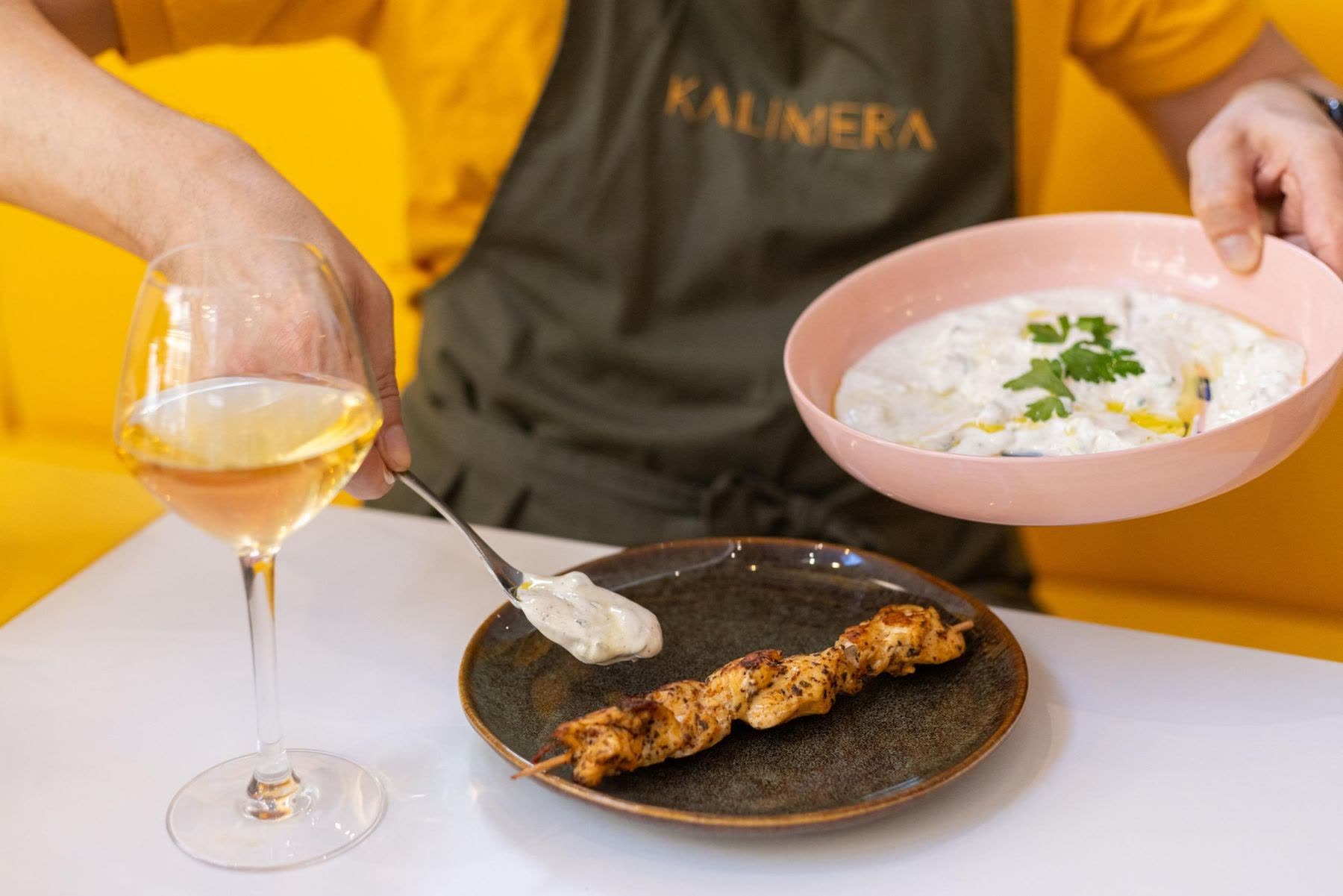 At a KALIMERA cooking workshop: a spoonful of tzatziki goes onto a plate beside a chicken skewer, with a bowl of tzatziki and a glass of white wine