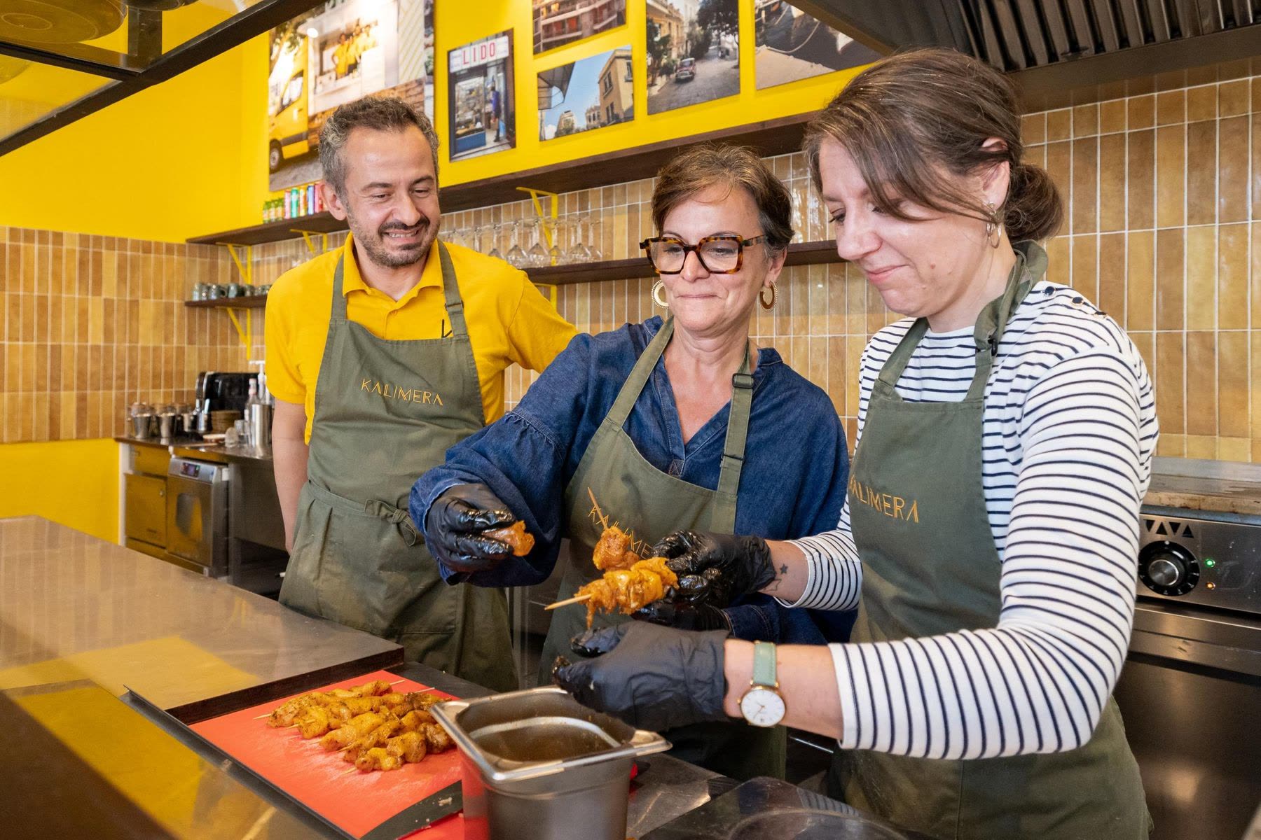 Un atelier de cuisine KALIMERA dans la cuisine de Bordeaux : deux personnes en tablier olive enfilent du poulet mariné sur des brochettes, sous l’œil amusé de leur hôte