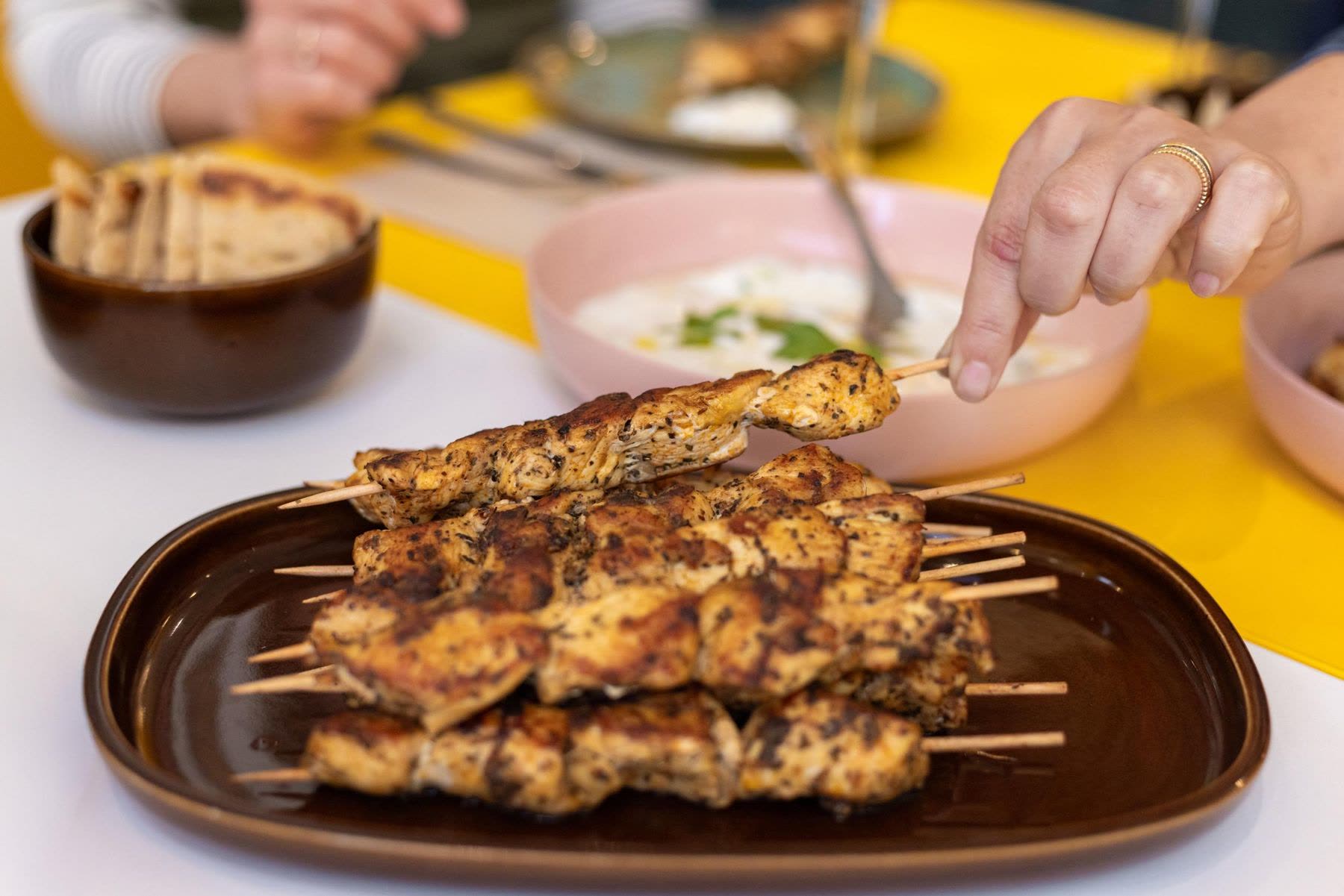 Chicken skewers on a brown plate after a KALIMERA cooking workshop, with tzatziki and pita behind, and a hand reaching for one