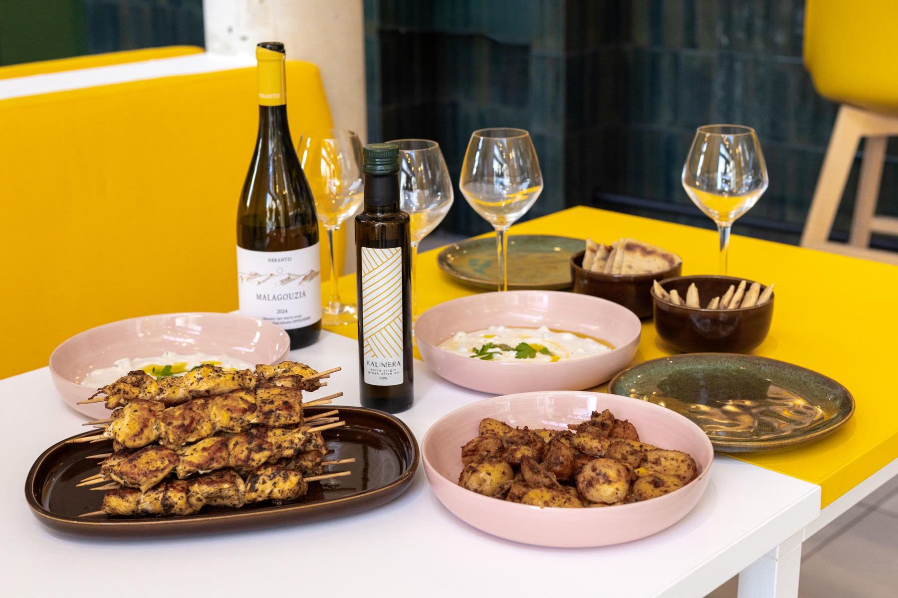 The table after a KALIMERA cooking workshop: chicken skewers, tzatziki, potatoes and pita, with a bottle of olive oil, a bottle of white wine and four glasses
