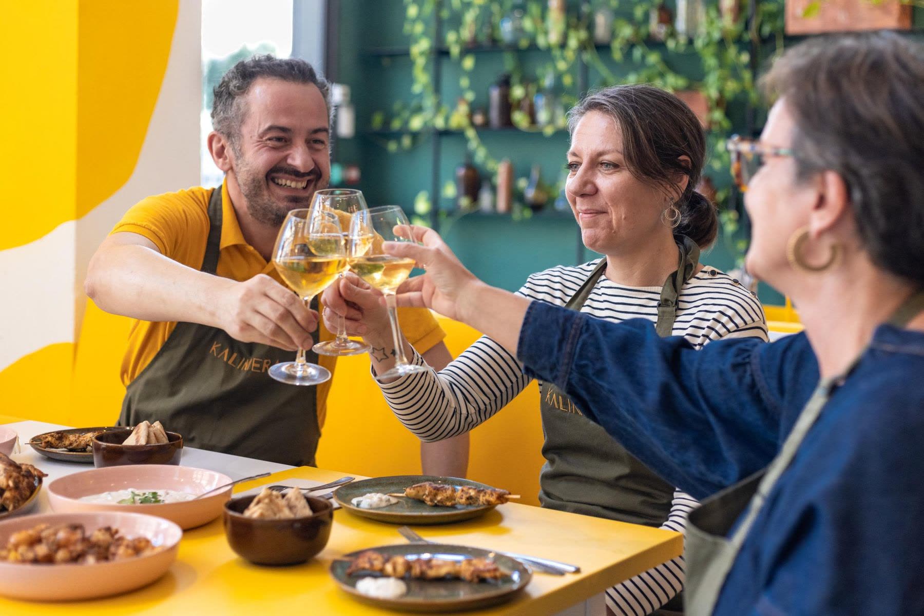 Après un atelier de cuisine KALIMERA à Bordeaux : l’hôte et deux convives trinquent au vin blanc au-dessus d’une table de brochettes de poulet, de tzatziki et de pita